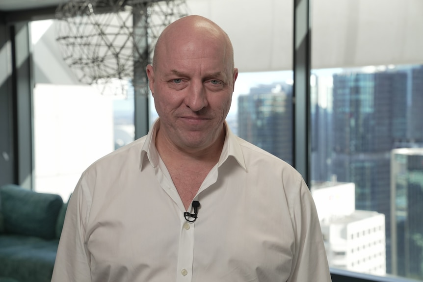 Matt smiles, is bald, caucasian, wears white shirt, background blurred, high rise buildings seen through glass window.