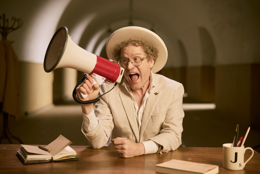 A man in a white hat and suit sits at a table yelling into a microphone