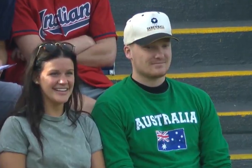 Baseball fan Jack Seiple sits in an Australia jersey next to a smiling woman in light green on stands.