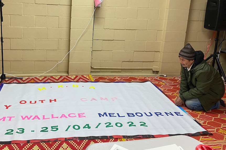An elderly man in a green jacket kneeling next to a large white banner which reads "MKBA Youth camp Mt Wallace Melbourne"
