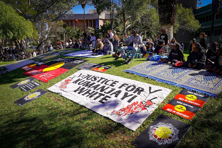 A large poster saying 'Justice for Kumanjayi White' laid on the lawn, with other protest posters and people surrounding.