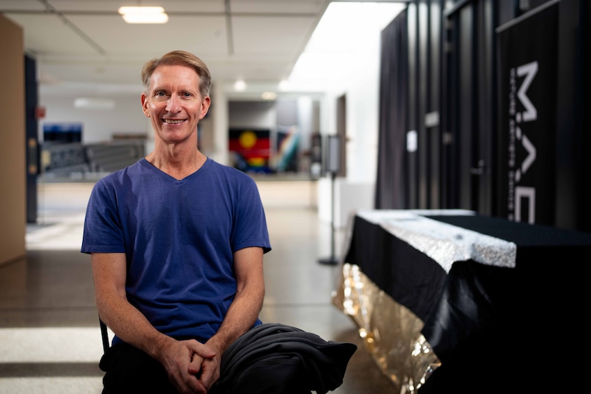 A man wearing a blue top is seated smiling for a photo