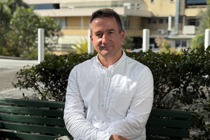 Man in white shirt sits on park bench with arms crossed