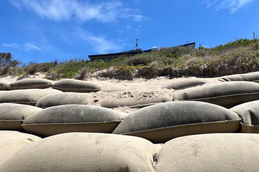 A building on top of a dune, at the foot of it are large sandbags. It is a sunny day.
