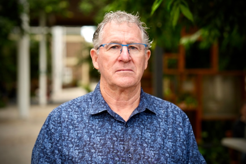A man with white hair with half-blue, half-clear rimmed glasses in a blue collared shirt stands outside, staring at the camera.