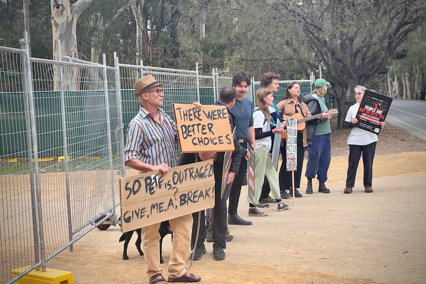 A line of protesters stand next to temporary fencing holding placards