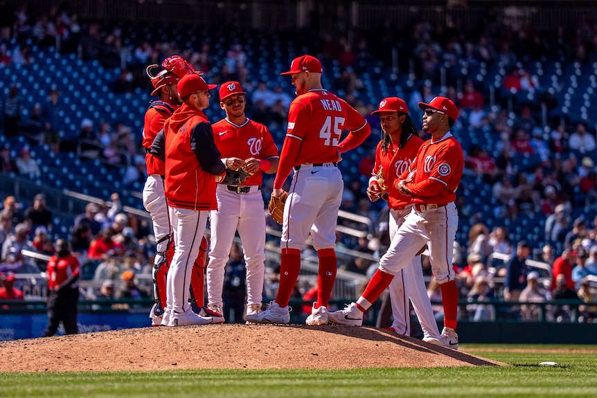 Curtis Mead stands on the mound with teammates, all wearing red, on the field during a baseball game.