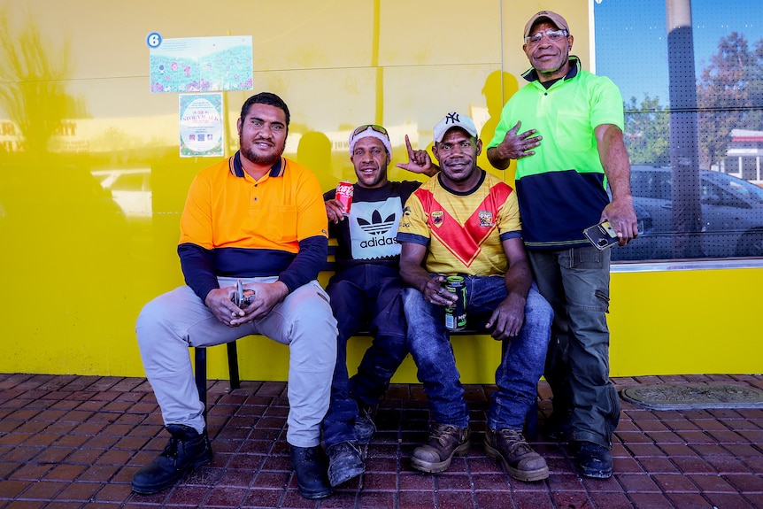 Four Pacific Islander workers wearing high-vis sit and stand against a yellow wall on a country street