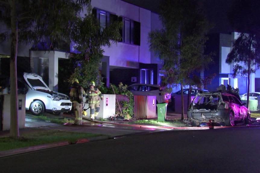 Two firefighters stand in the driveway of a row of townhouses between a burnt out car and a damaged car at night.