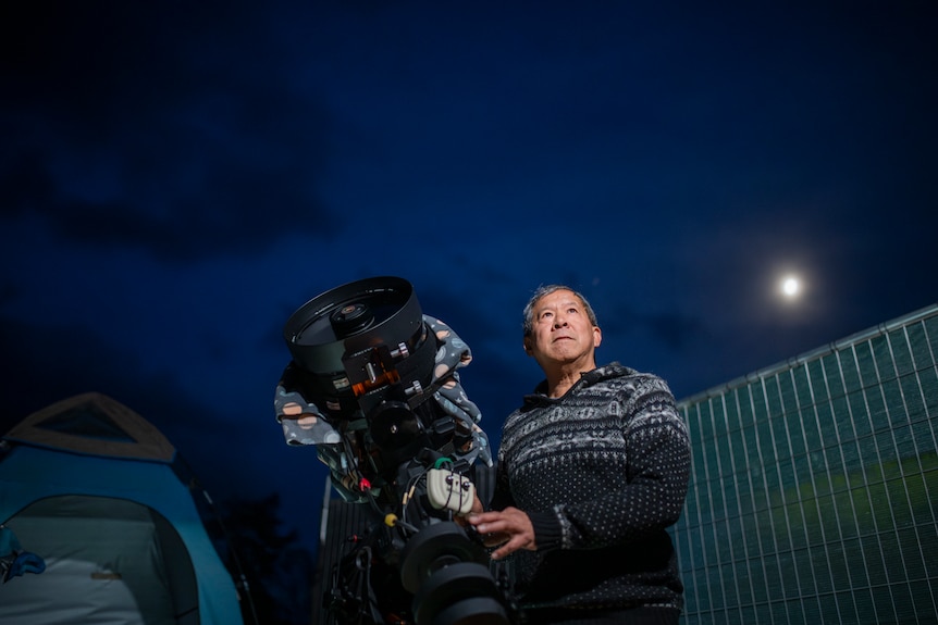 A man in a black and white patterned jumper stands next to a telescope the same size as him under an electric blue sky.