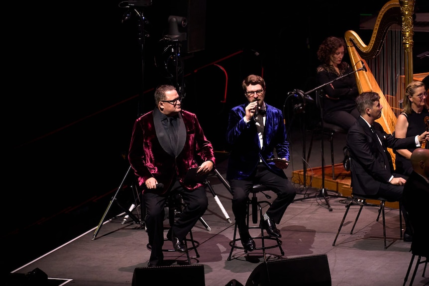 Andrew Pogson and Dan Golding in velvet jackets speak on stools beside an onstage orchestra, with harp and strings.