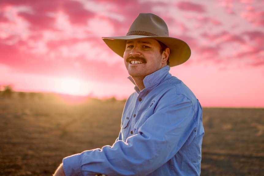 Man with akubra looks at camera, pink sky and field in background.