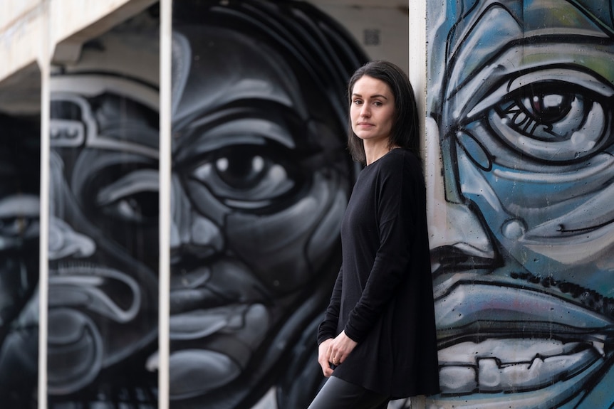 Emily Corner, who has dark brown hair and is wearing black, poses for a photo in front of a moody mural