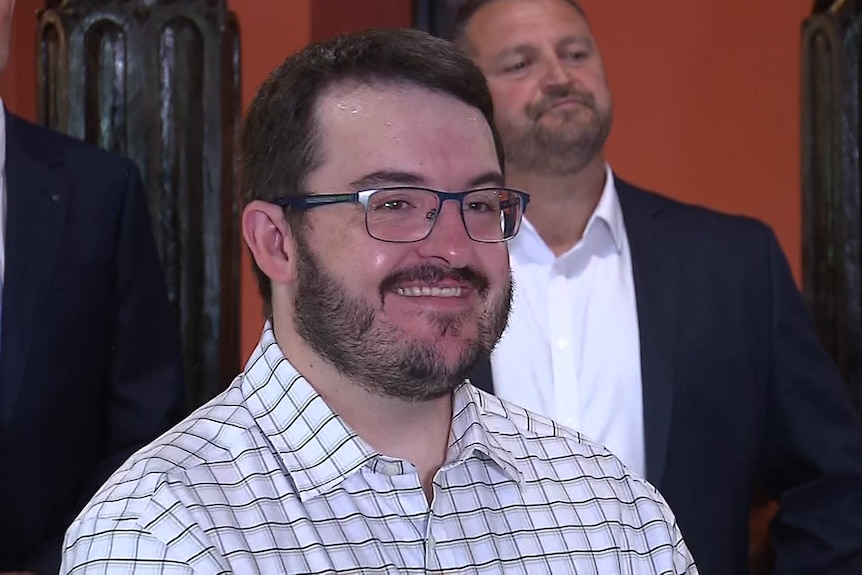 Jason Virgo in a business shirt smiles and stands in front of other One Nation MPs