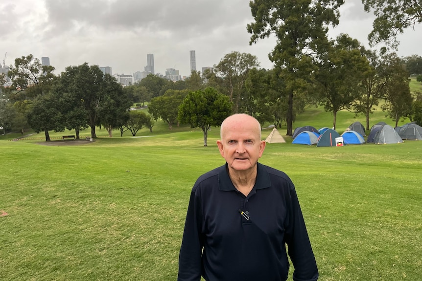 A man stands in front of a grassy areas with tents in the background.