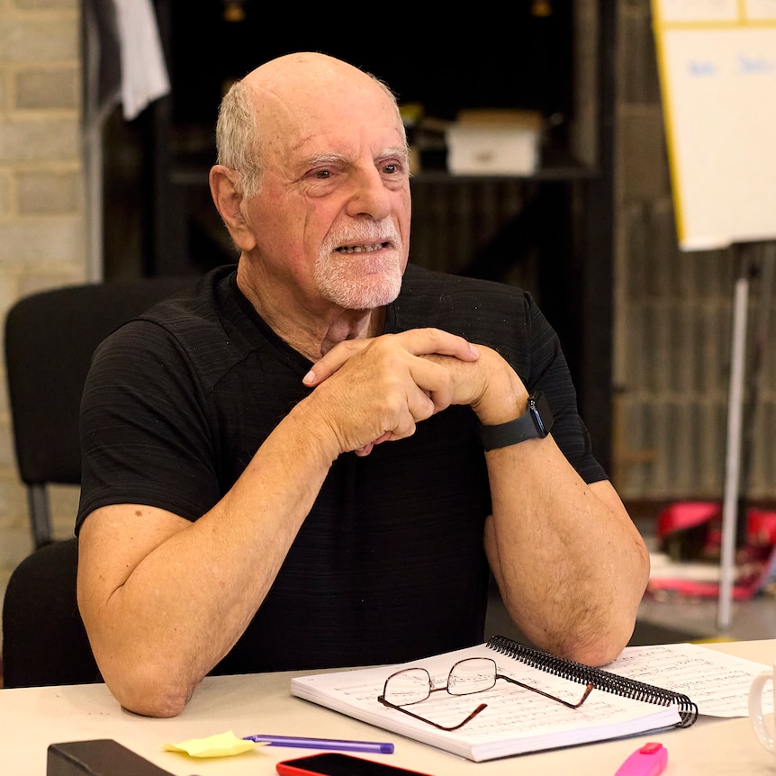 George Palmer sits at a desk in a black t-shirt with a serious expression. There is a score and glasses in front of him.