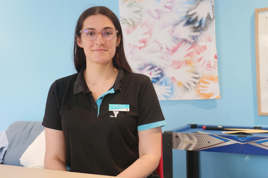 A woman sitting down wearing a black collared shirt with a nametag and The Y logo, in front of a wall with student made artwork