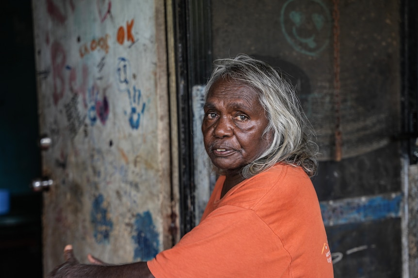 An Aboriginal woman wearing an orange top and grey hair looking at camera