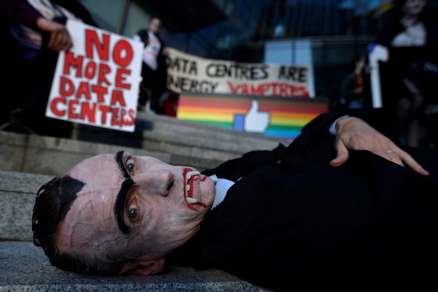 A man in a vampire outfit lies on the steps of an office building with protest signs against data centres behind him.