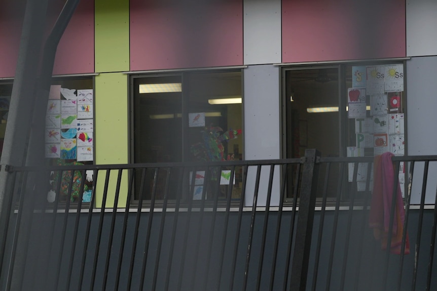 Children's artwork lines the windows of a classroom seen through the metal bars of a fence.