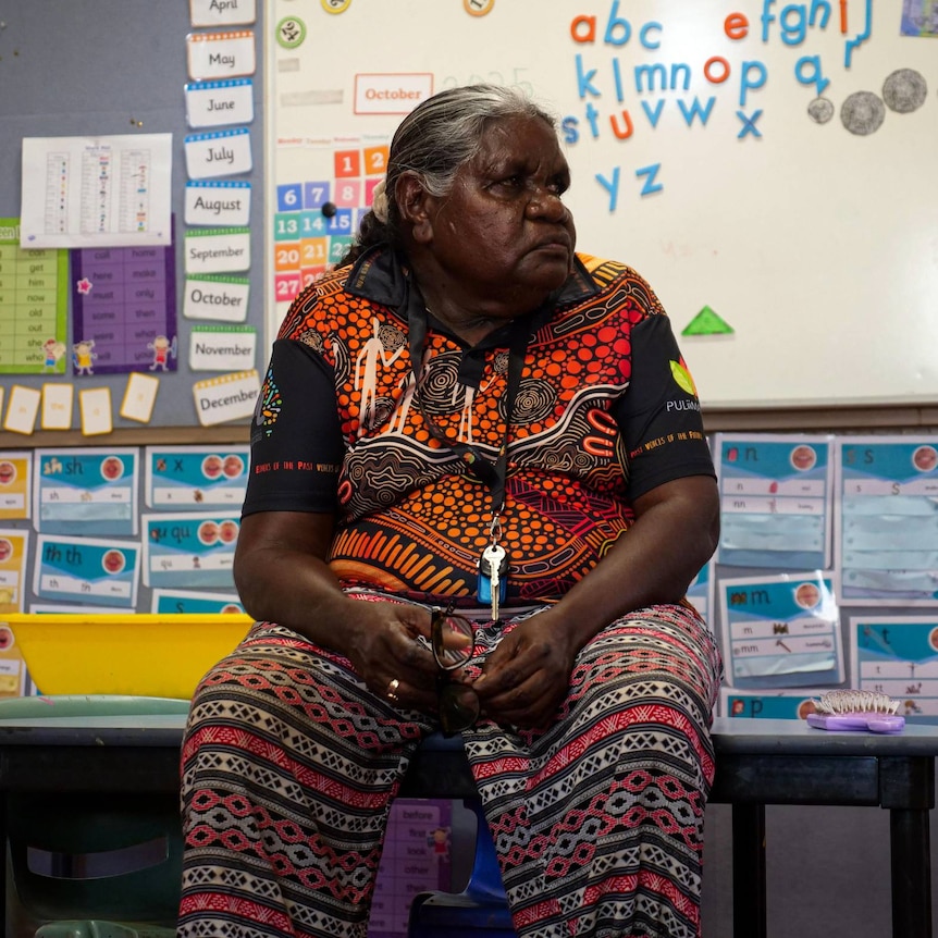 An older Aboriginal woman with silver hair in a bun sits on a desk in a classroom.
