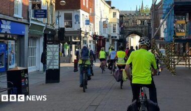 A group of cyclists wearing hi vis and helmets cycling through Salisbury high street with shops either side of them, some large scaffolding and a large stone arch - High Street Gate - and spires in the background.