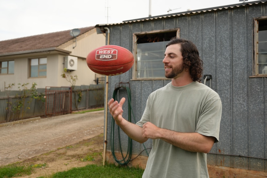 A man with long dark hair tosses a football in the air to himself.