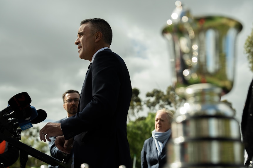 Side view of Peter Malinauskas speaking behind a group of media microphones while standing near a large trophy