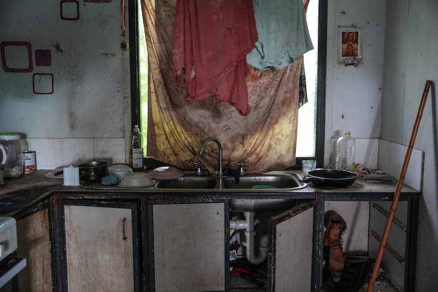 A kitchen sink with broken cupboards underneath and a window above covered by dirty sheet