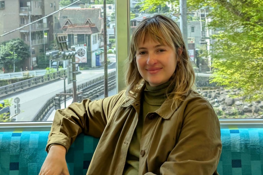A woman wearing a brown coat smiles from a blue seat.