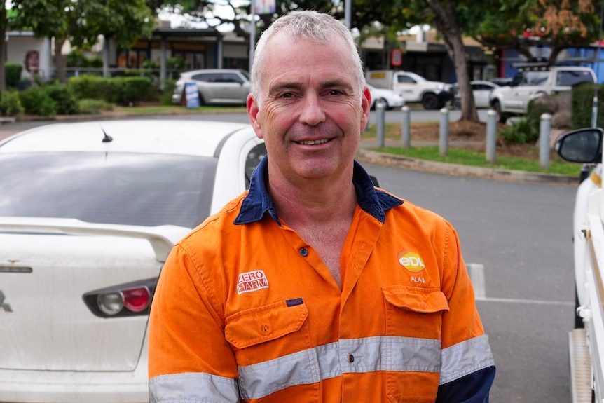 A smiling, middle-aged man with grey hair wears a high-vis work shirt while standing in a car park.