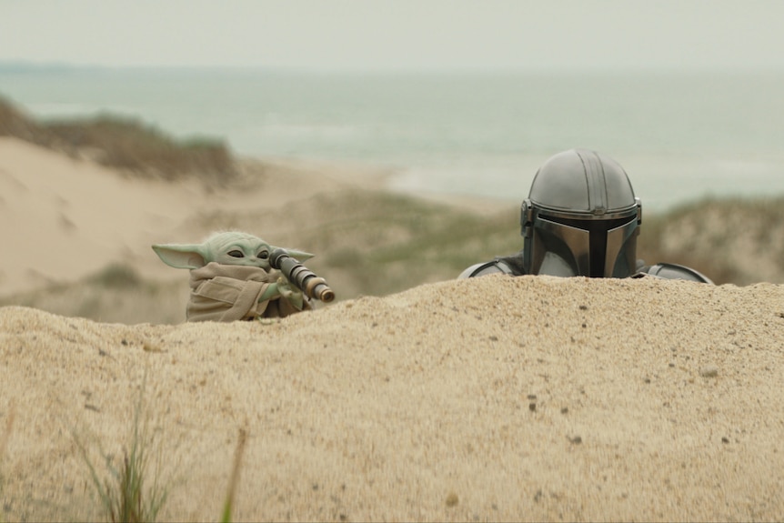 A small creature and a helmeted man peek out from behind a sand dune.