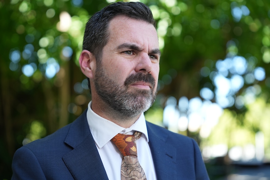 A man with short dark hair and a beard wearing a dark suit looks to the right