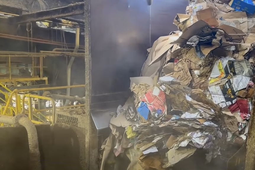 A pile of cardboard inside a steel container near a set of stairs with yellow handrailing.