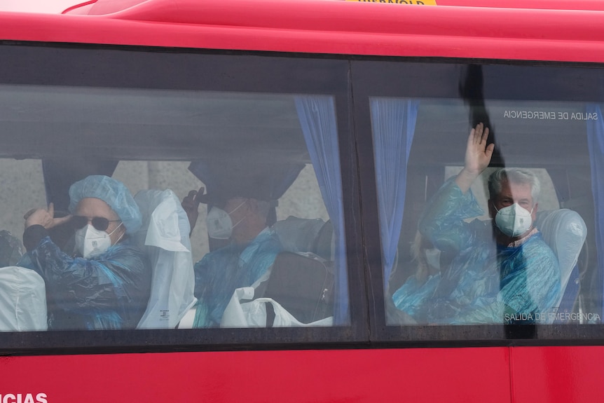 People in blue hazmat suits on board a bus wave as they go to the airport to fly home.