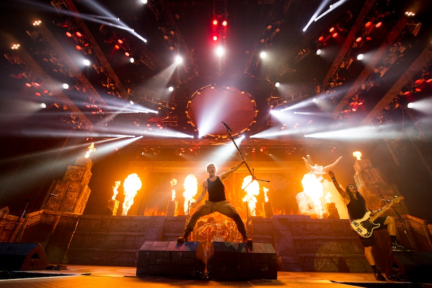 Iron Maiden performs on stage with bright lights, flames, and a raised microphone stand at center stage