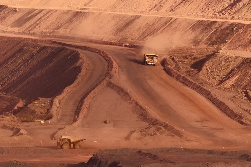 A wide shot of a large mine site with red dirt slopes, two mining trucks appear tiny on the landscape.