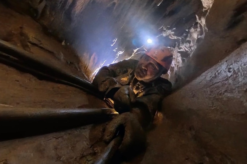 A man in an orange helmed climbs down a narrow cave, with the only light coming from headlamps.