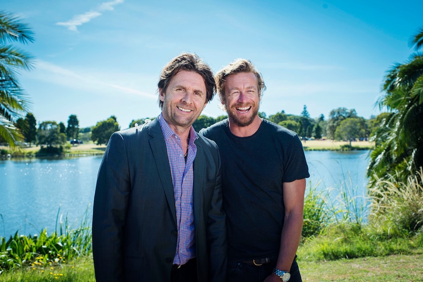 Two men stand together and smile with a lake in the background.