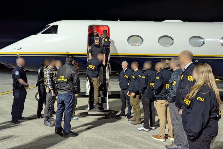 A man walking down the stairs of a plane, surrounded by many armed FBI agents.