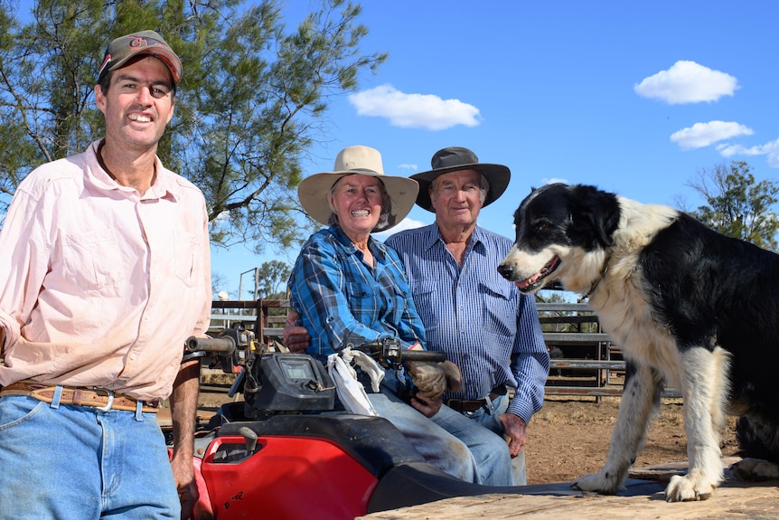 A dog and three farmers all lean on a quad bike on a farm.