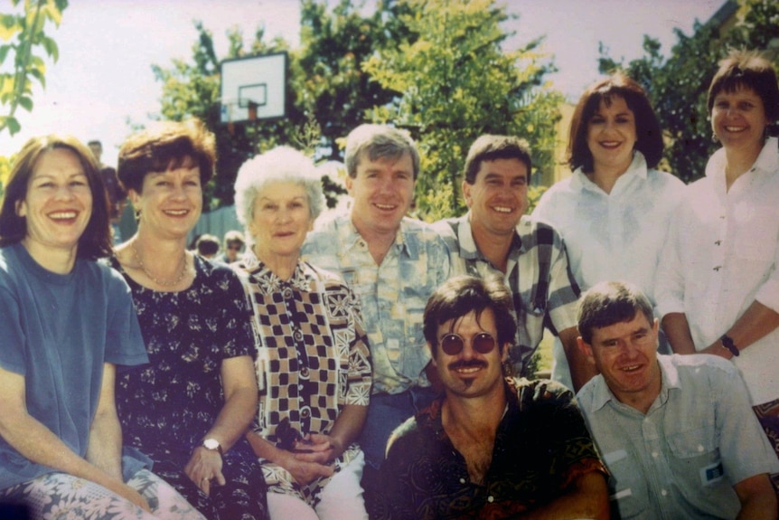 A photograph of an older woman surrounded by eight middle-aged people - four men and four women.