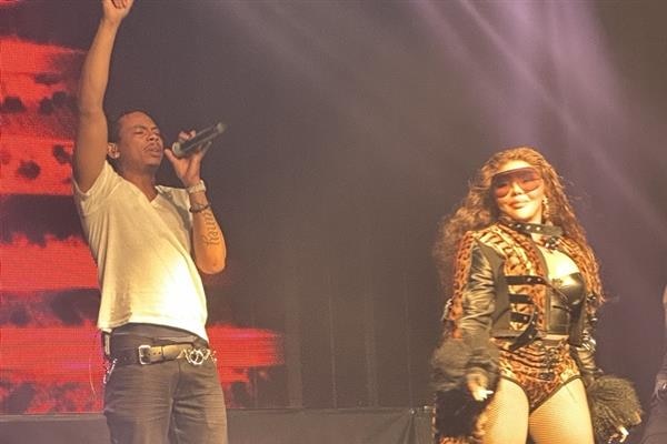 Tayy Brown with arm raised singing into a mic, Lil' Kim looking on at the audience.