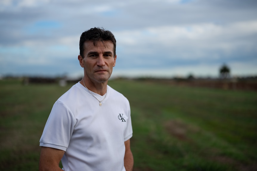 A man in his 40s with dark cropped hair wearing a white t-shirt stands in a green paddock on a farm.
