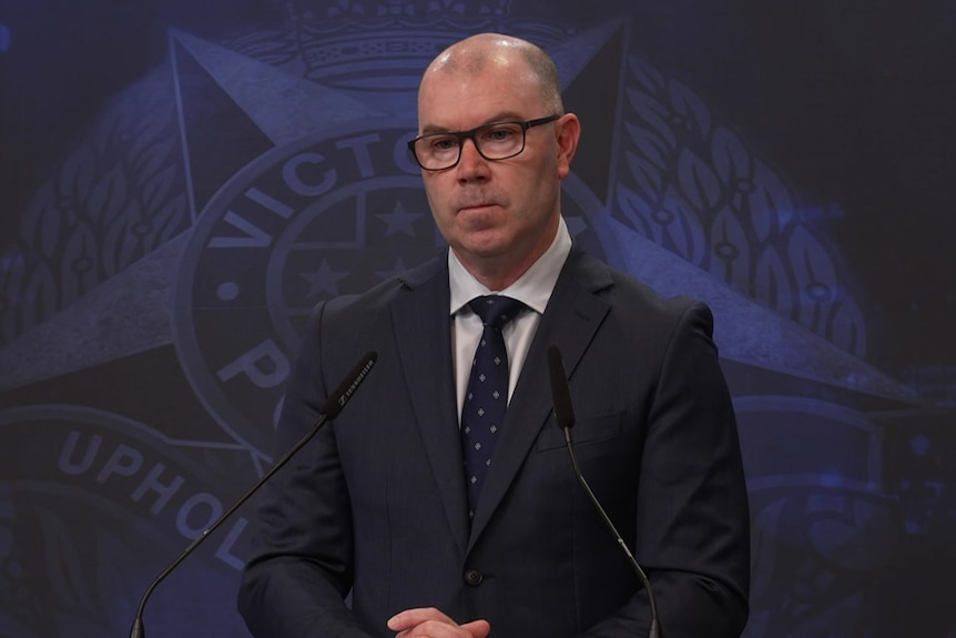 A man in a suit with glasses stands at a podium addressing media.
