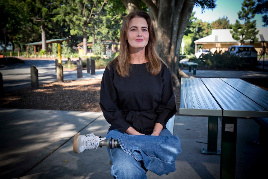 A white woman with long brown hair and a prosthetic leg sitting on a park bench
