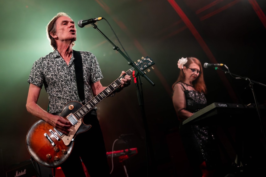 Mick Medew holds a guitar and sings into a microphone while Ursula Collie plays a keyboard.