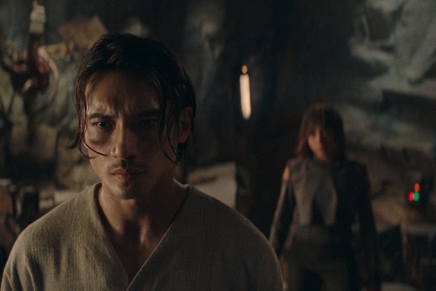 A scene in a science fiction TV series, featuring a man standing in a cave with a woman looking at him in the background.