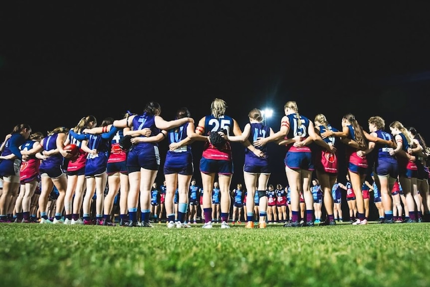 A women's AFL team stand in a pre-game huddle, each players is wearing two red bands on their left arm.