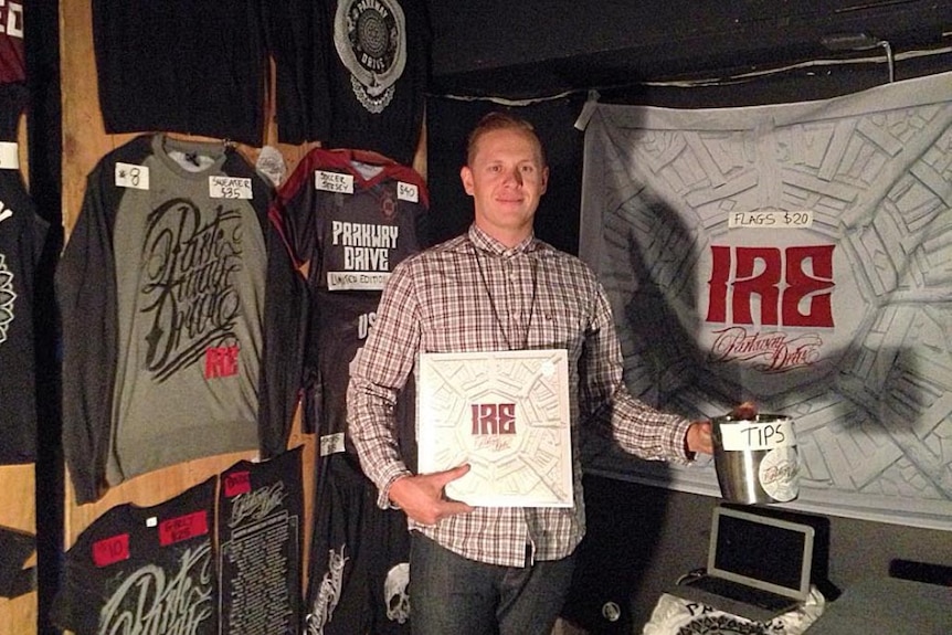 Blonde man in checked shirt holding Parkway Drive album Ire with merchandise on the walls around him